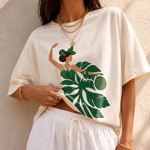 May include: An ivory short-sleeved t-shirt featuring a graphic print of a figure with dark hair adorned with green leaves, wearing a skirt made of large green monstera leaves and holding a textured green fruit. The shirt is labeled Ivory CC1717.