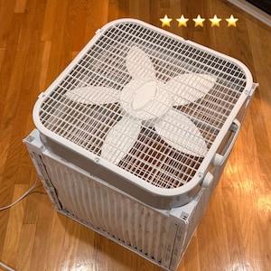 May include: A white box fan with a square design, featuring a protective grid and a set of five fan blades. The fan sits atop a rectangular air filter. The fan is designed to circulate air and improve air quality.