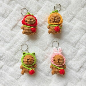 May include: Four crocheted capybara keychains, each featuring a brown body and a unique hood: red, orange, green frog-themed, and pink rabbit-eared. Each capybara wears a green strap with a small red strawberry charm attached to the side.