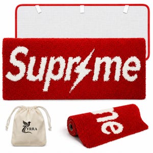 May include: A rectangular red rug with a white tufted design featuring the text "Supr me" with a lightning bolt replacing the letter "e". The rug has a white non-slip backing with a red border. A small beige drawstring bag with a black bird logo is included.