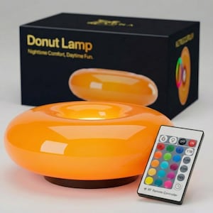 Puede incluir: Una lámpara naranja con forma de donut y un mando a distancia. La caja tiene el texto "Donut Lamp Nighttime Comfort, Daytime Fun". El mando a distancia tiene botones para la selección de color y otras funciones.