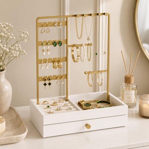 May include: A gold metal jewelry stand with a white wooden base featuring a drawer. The stand includes multiple hooks for earrings, necklaces, and bracelets. The white base contains compartments for rings and watches, organized with various pieces of jewelry.