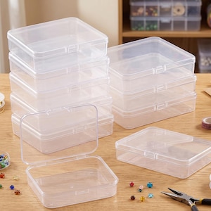 May include: Clear plastic storage boxes with hinged lids are stacked on a wooden surface. Several boxes are open, revealing their interior. The boxes are rectangular and appear to be empty, suitable for organizing small items like beads or craft supplies.