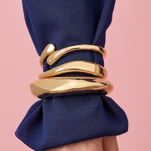 May include: Three gold-colored, chunky bracelets are stacked on a navy blue sleeve against a pink background. The bracelets have a smooth, polished finish and a modern design. The bracelets are wrapped around the sleeve.