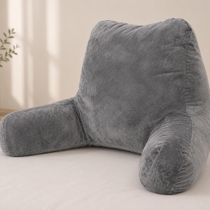 May include: A dark gray reading pillow with a plush, velvet-like texture. This backrest cushion features a tall, upright back section and two integrated side arms, providing a structured shape designed to support an upright seated position.