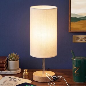 May include: A cylindrical table lamp with a white fabric shade and a wooden base. The lamp is turned on, emitting a soft glow. The lamp is on a wooden desk with a succulent plant and a green mug.