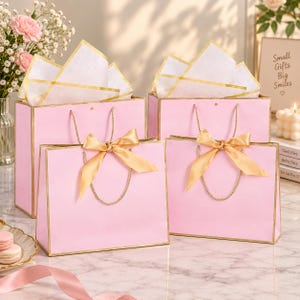 May include: Four light pink gift bags with gold trim and gold rope handles. Each bag features a gold satin ribbon bow tied at the center. Two bags are taller and filled with white tissue paper, while two are shorter and positioned in the front.
