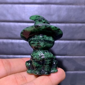 May include: A small, carved figurine of a frog wearing a pointed hat, crafted from a dark green stone with black speckled inclusions. The polished mineral sculpture features a smooth texture and distinct natural patterns throughout the material.
