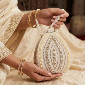 Può includere: Una pochette a forma di goccia con intricate perline bianche e dorate, ricami metallici e una struttura rigida. La borsa include un manico di perline ed è decorata con file di piccole perle e dettagli dorati riflettenti.