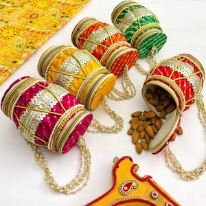 Puede incluir: Una colección de coloridos recipientes cilíndricos de tela con asas de cuentas. Los recipientes son en tonos de rojo, naranja, amarillo y verde, con detalles plateados y dorados. Un recipiente está abierto, derramando almendras. Un tejido amarillo estampado está en el fondo.