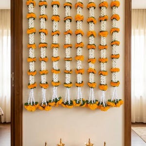 Puede incluir: Ocho guirnaldas verticales de flores de caléndula blancas y naranjas alternadas cuelgan de una barra horizontal de latón con campanillas. Debajo, cuatro lámparas de aceite de latón y una pequeña mesa de madera con un cuenco de pétalos.