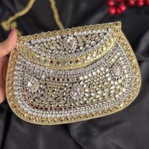 Peut inclure: Une pochette or et argent ornée d'une chaîne. La pochette est recouverte d'un motif de pierres transparentes et argentées, avec une bordure dorée. Le sac est tenu par une personne avec du vernis à ongles rose.