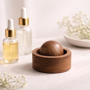 Puede incluir: Una bola de masaje de madera descansa en un cuenco de madera a juego. Dos frascos cuentagotas de vidrio con tapas doradas están en el fondo. Uno contiene un líquido amarillo, el otro es transparente. Hay flores blancas.