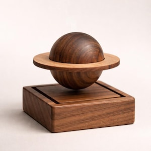 Op de afbeelding: Een houten sculptuur in de vorm van een planeet, met een donkerbruine bol, een lichtbruine ring en een vierkante basis. De bol lijkt boven de ring te zweven, die bovenop de basis staat. De sculptuur is gemaakt van hout met een natuurlijke afwerking.