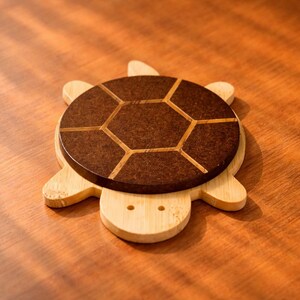 May include: A wooden turtle-shaped coaster with a dark brown, geometric patterned top. The coaster is made of light-colored wood and sits on a wooden surface. The design is a decorative and functional item for home decor.