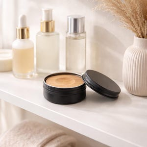 Op de afbeelding: Een zwarte metalen container met een houten inzetstuk en een los deksel staat op een witte plank. Op de achtergrond staan ​​verschillende heldere glazen flessen met gouden en zilveren doppen, samen met een witte vaas met gedroogde planten. De scène suggereert een spa- of huidverzorgingsthema.