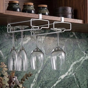 May include: A white metal under-cabinet wine glass rack with four clear glass stems hanging upside down. The rack features a rectangular frame design that slides onto a shelf, providing storage for stemware in a kitchen setting.