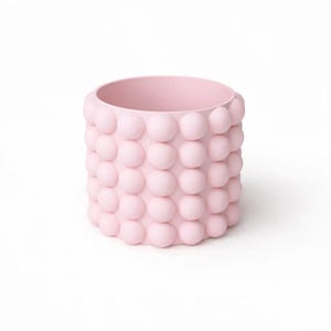 May include: A cylindrical light pink planter featuring a textured exterior surface covered in a repeating pattern of raised, spherical bubble-like protrusions arranged in uniform vertical and horizontal rows around the entire vessel.