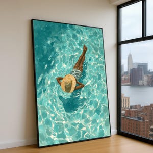 Peut inclure: Une impression d'art encadrée représente une personne flottant dans une piscine turquoise, portant un chapeau de paille et un maillot de bain à motifs. L'œuvre est exposée à l'intérieur, adossée à un mur blanc, avec vue sur la ville par une fenêtre.