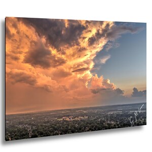 May include: A landscape photograph featuring a dramatic sunset sky filled with large, golden-orange and dark grey storm clouds hovering over a sprawling suburban area with dense green trees and a signature in the bottom right corner.