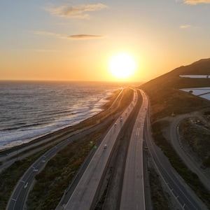 May include: An aerial view of a multi-lane highway running parallel to the ocean coastline during a golden sunset. The sun is low on the horizon, casting a warm glow over the dark water, the asphalt road, and the white agricultural structures.