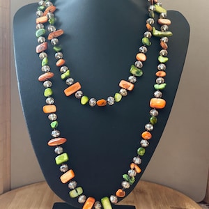Könnte beinhalten: Eine mehrreihige Halskette, präsentiert auf einer schwarzen Büste. Die Kette besteht aus orangefarbenen, grünen und transparenten Perlen. Die unregelmäßig geformten Perlen sind aufgefädelt und bilden ein einzigartiges, farbenfrohes Accessoire.