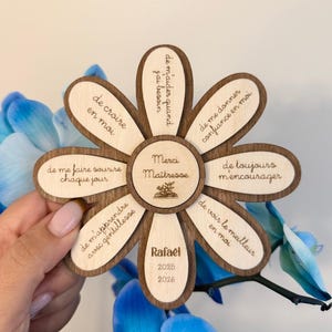 Peut inclure: Un cadeau en forme de fleur en bois avec une bordure marron foncé et des pétales en bois clair. Le centre dit "Merci Maîtresse" et "Rafael 2025 2026". Chaque pétale porte une phrase en français. Le cadeau est tenu devant une fleur bleue.