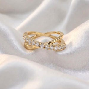 May include: A gold-colored ring with a crisscross design, set with small, clear gemstones. The ring is displayed on a white, silky fabric, highlighting its elegant and delicate features. The ring's design is modern and sophisticated.