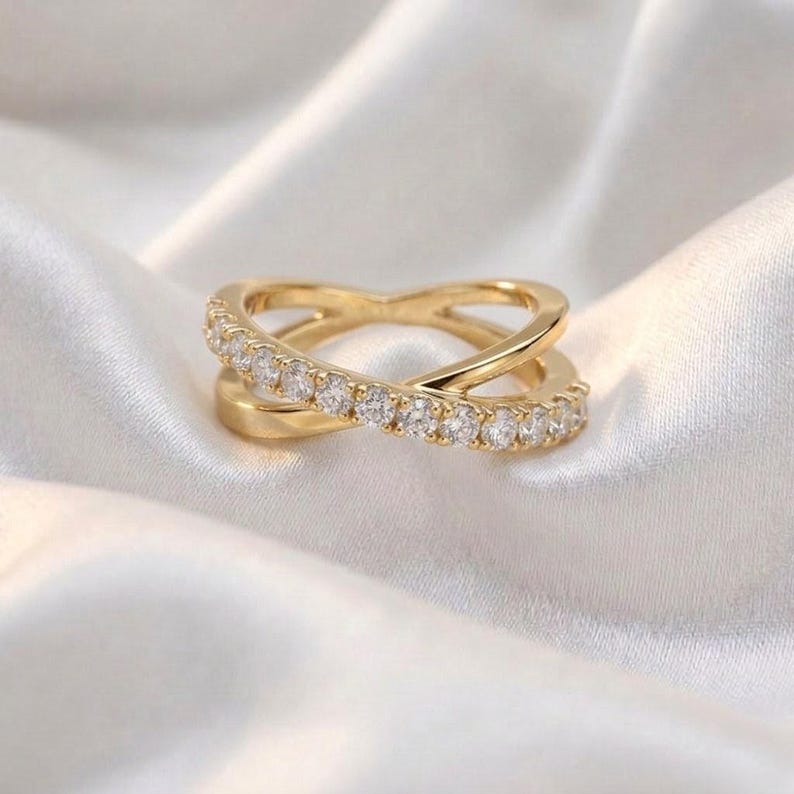 May include: A gold-colored ring with a crisscross design, set with small, clear gemstones. The ring is displayed on a white, silky fabric, highlighting its elegant and delicate features. The ring's design is modern and sophisticated.