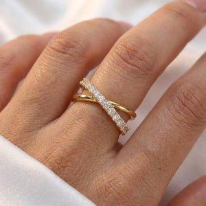 May include: A gold-colored criss-cross ring with a row of small, clear stones on one band. The ring is worn on a finger, and the background is a soft, neutral color.