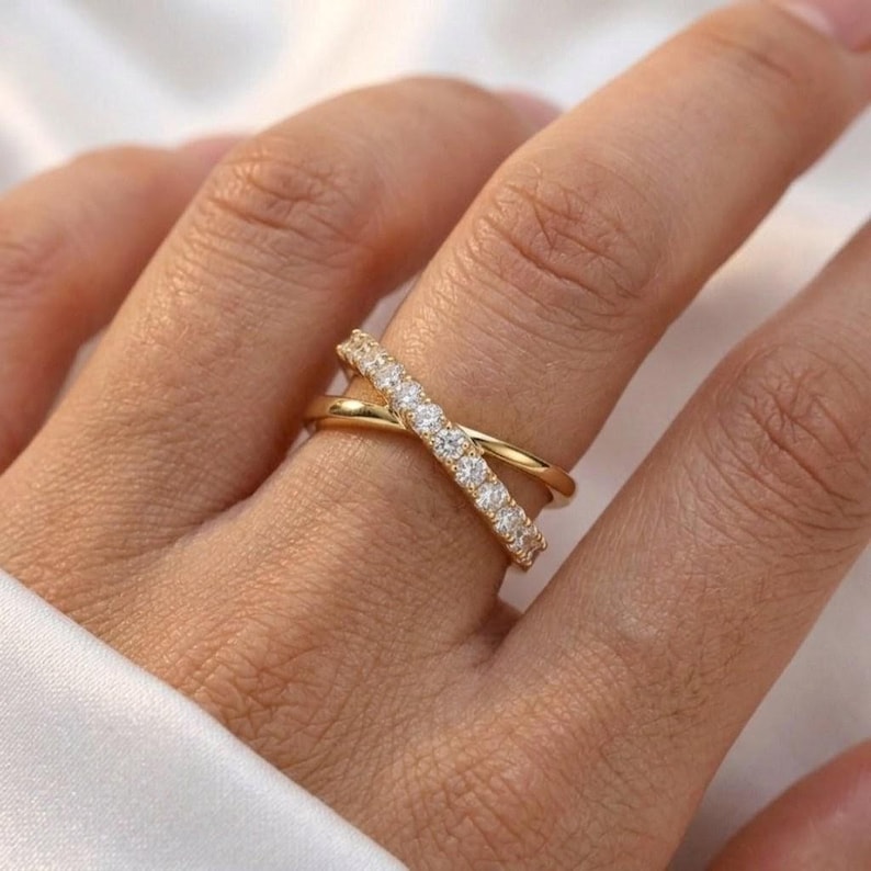 May include: A gold-colored criss-cross ring with a row of small, clear stones on one band. The ring is worn on a finger, and the background is a soft, neutral color.