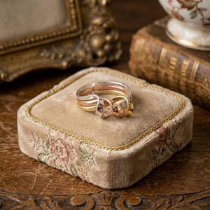 May include: A multi-tone ring with silver, rose gold, and yellow gold bands. The ring is displayed on a square, velvet jewelry box with floral embroidery and a gold trim. The ring's design features a knot-like detail.