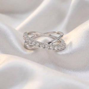 May include: A silver-colored ring with a crisscross design. One band is encrusted with small, clear gemstones. The ring is set against a white, silky background, creating a contrast that highlights the ring's details.