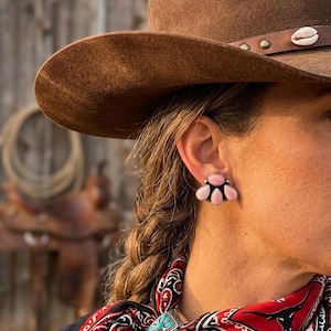 May include: A close-up of a person wearing a brown cowboy hat and pink gemstone earrings. The earrings are clustered in a floral design. The person is also wearing a red and black patterned scarf with a turquoise brooch.