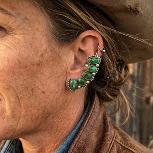 Op de afbeelding: Een oormanchet met vijf ovaalvormige groene stenen met donkere aders, gezet in een zilverkleurig metalen frame met een kralenrand. Het sieraad volgt de welving van het oor, aangevuld met een kleine zilveren ring erboven.