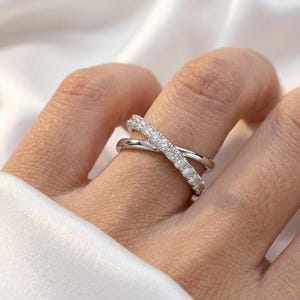 May include: A silver-toned criss-cross ring with a row of small, clear stones set into one band. The ring is worn on a finger, with a soft white fabric in the background. The ring's design creates an X shape.