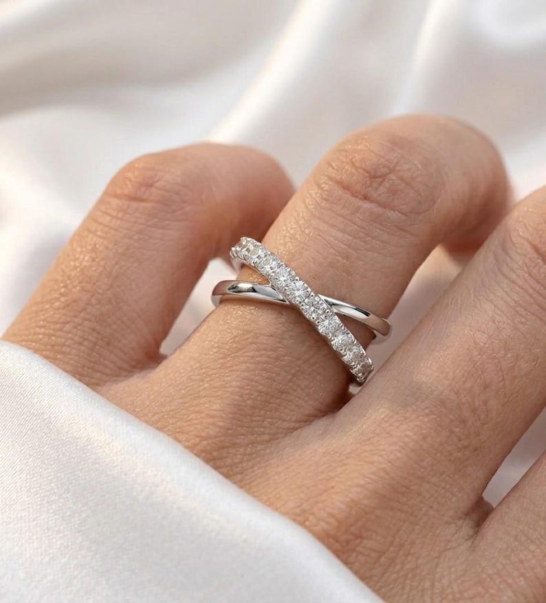 May include: A silver-toned criss-cross ring with a row of small, clear stones set into one band. The ring is worn on a finger, with a soft white fabric in the background. The ring's design creates an X shape.
