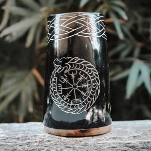Peut inclure: Une corne à boire noire avec un motif gravé blanc. La corne présente un motif de nœud celtique en haut et un symbole circulaire avec un serpent et des runes au centre. La base de la corne est de couleur marron clair.