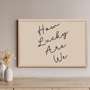 Könnte beinhalten: Gerahmter Druck mit dem Text "How Lucky Are We" in schwarzer Schrift auf beigem Hintergrund. Der Rahmen ist hellbraun. Der Druck wird auf einer Holzoberfläche präsentiert.