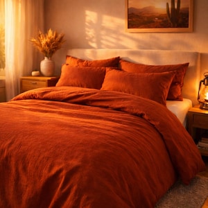 May include: A bed with a rust-colored duvet cover and matching pillowcases. The bedding is arranged on a bed with a white headboard. A wooden nightstand with a lamp and a vase of dried flowers is visible.