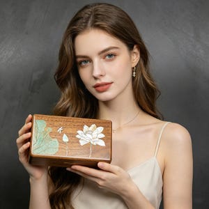 以下が含まれることがあります： 蓮の花のデザインが施された長方形の木製ボックス。ボックスは茶色で、前面に白と緑の花柄があります。ボックスは人の前に保持されています。