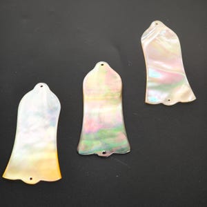 May include: Three iridescent, bell-shaped guitar headstock inlays. The inlays have a pearlescent finish with hues of white, yellow, pink, and green. Each inlay has a small hole at the top and bottom for attachment. The background is a solid black.