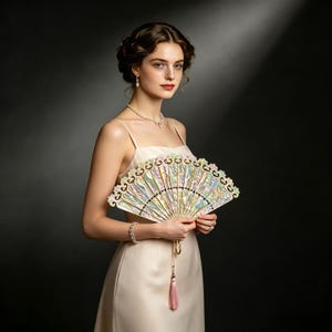 May include: A handheld folding fan featuring an iridescent, mother-of-pearl style finish with intricate cut-out patterns along the top edge. The fan includes a delicate pink tassel hanging from the base, held in a neutral-toned, spaghetti-strap dress.