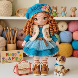 Könnte beinhalten: Eine handgefertigte Häkelpuppe mit roten Locken, die einen blauen Barett mit Blumen, ein blaues Kleid und eine cremefarbene Weste trägt. Die Puppe hat eine passende Handtasche und Stiefel. Eine Häkelkatze mit blauem Band sitzt daneben. Die Puppe ist mit "Elena & Pip" beschriftet.