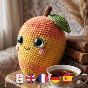 May include: A handmade crochet peach with large, expressive eyes, rosy cheeks, and a smiling mouth. The peach is yellow and orange, with a green leaf and brown stem. A cup of tea sits nearby, with flags of different countries.