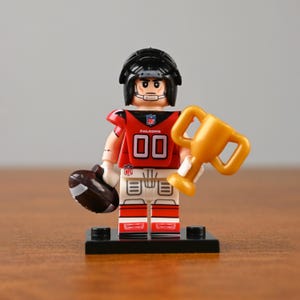 May include: A miniature football player figurine wearing a red and black uniform with the number 00. The figurine is holding a brown football in one hand and a gold trophy in the other. The figure is on a black base.