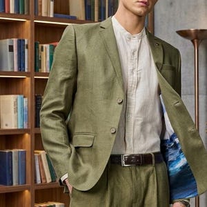 Op de afbeelding: Een olijfgroene blazer en bijpassende broek gecombineerd met een lichtbeige overhemd. De blazer heeft twee knopen aan de voorzijde, klepzakken en een blauw-witte voering met patroon die zichtbaar is aan de binnenkant van het open jasje.