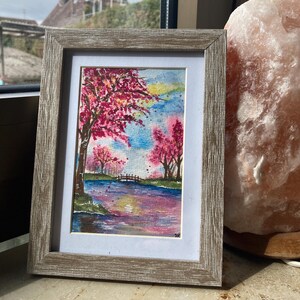 Könnte beinhalten: Ein gerahmtes Aquarellgemälde einer Landschaftsszene. Das Kunstwerk zeigt einen Fluss mit einer kleinen Brücke, Bäume mit rosa Blüten und einen farbenfrohen Himmel. Der Rahmen ist hellbraun.