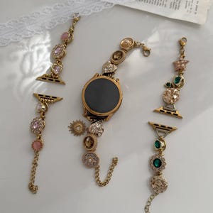 May include: A gold-toned smartwatch with a circular black face, featuring a decorative watch band composed of multiple gold-colored chain links, charms, and embedded crystals in shades of pink, brown, green, and clear, arranged in a multi-piece set.