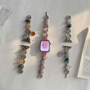 May include: Silver Apple Watch with a pink heart on the screen, and decorative bands. The bands feature colourful beads and charms, including pink, yellow, and blue accents. The time displayed is 17:25.